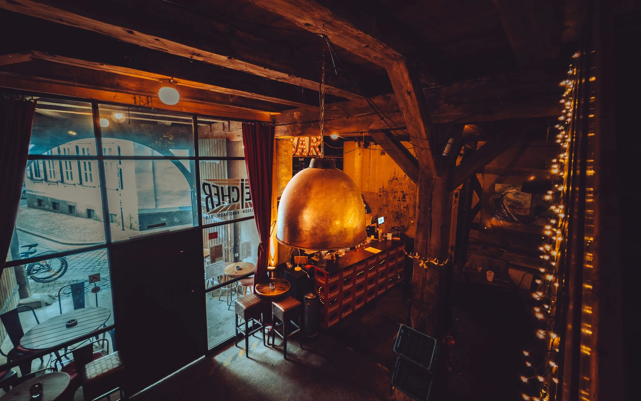 Innenraum eines gemütlichen Café oder Bars mit warmen Lichteffekten, Holzbalken und kleinen Tischen.