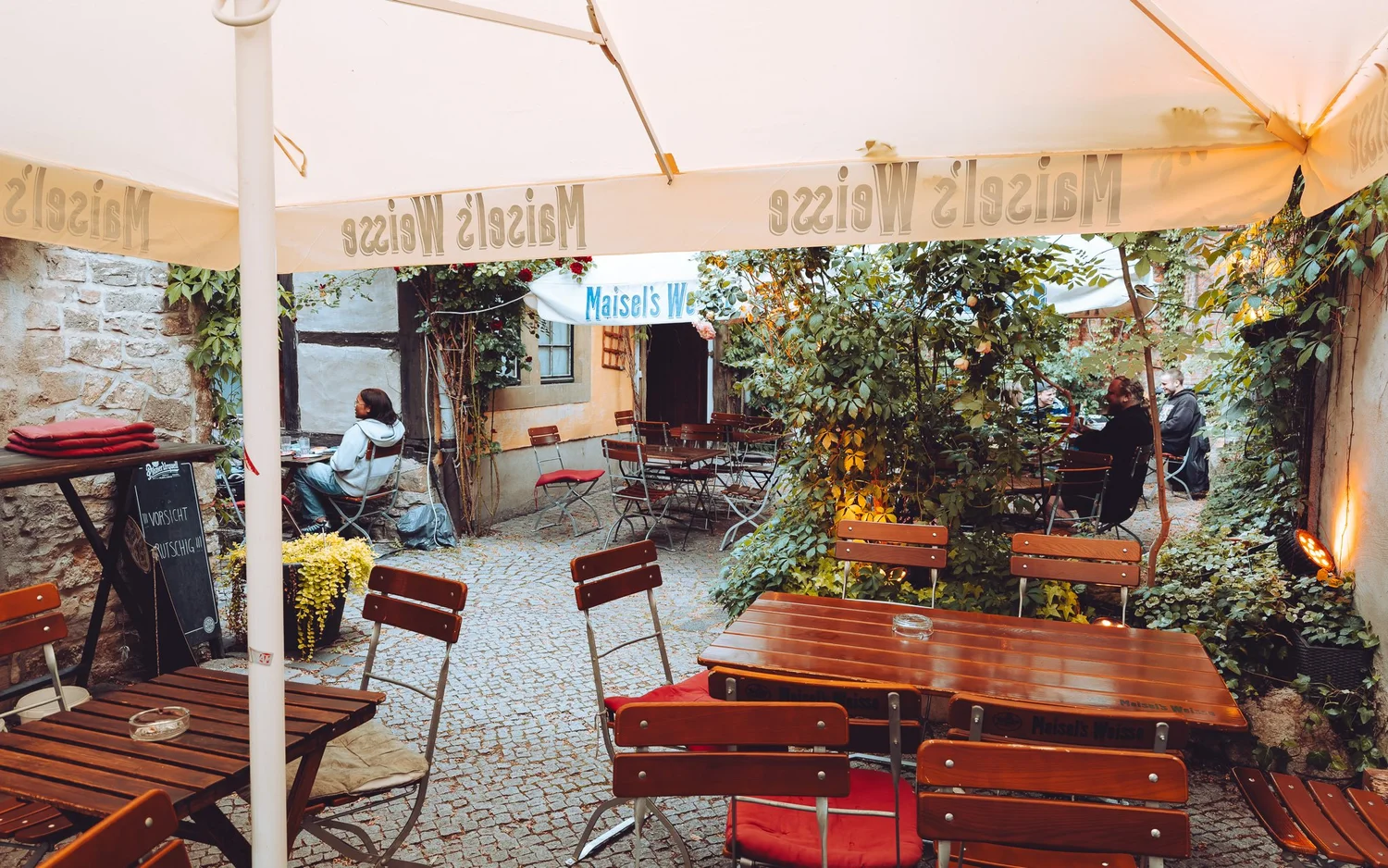 Leerer Biergarten-Außenbereich mit Tischen, Stühlen und umgebenden Pflanzen unter einem großen Sonnenschirm